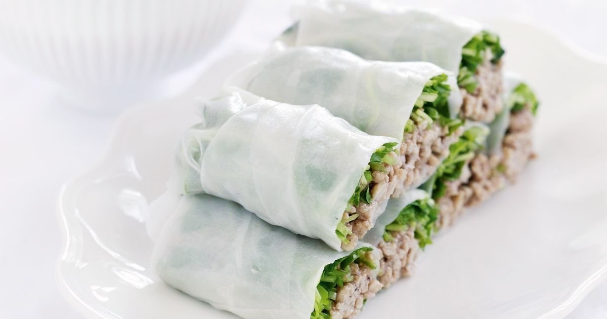 Four Vietnamese spring rolls filled with greens and minced meat are arranged neatly on a white plate. The thin rice paper wrapping reveals the fresh ingredients inside. A blurred white bowl is in the background.